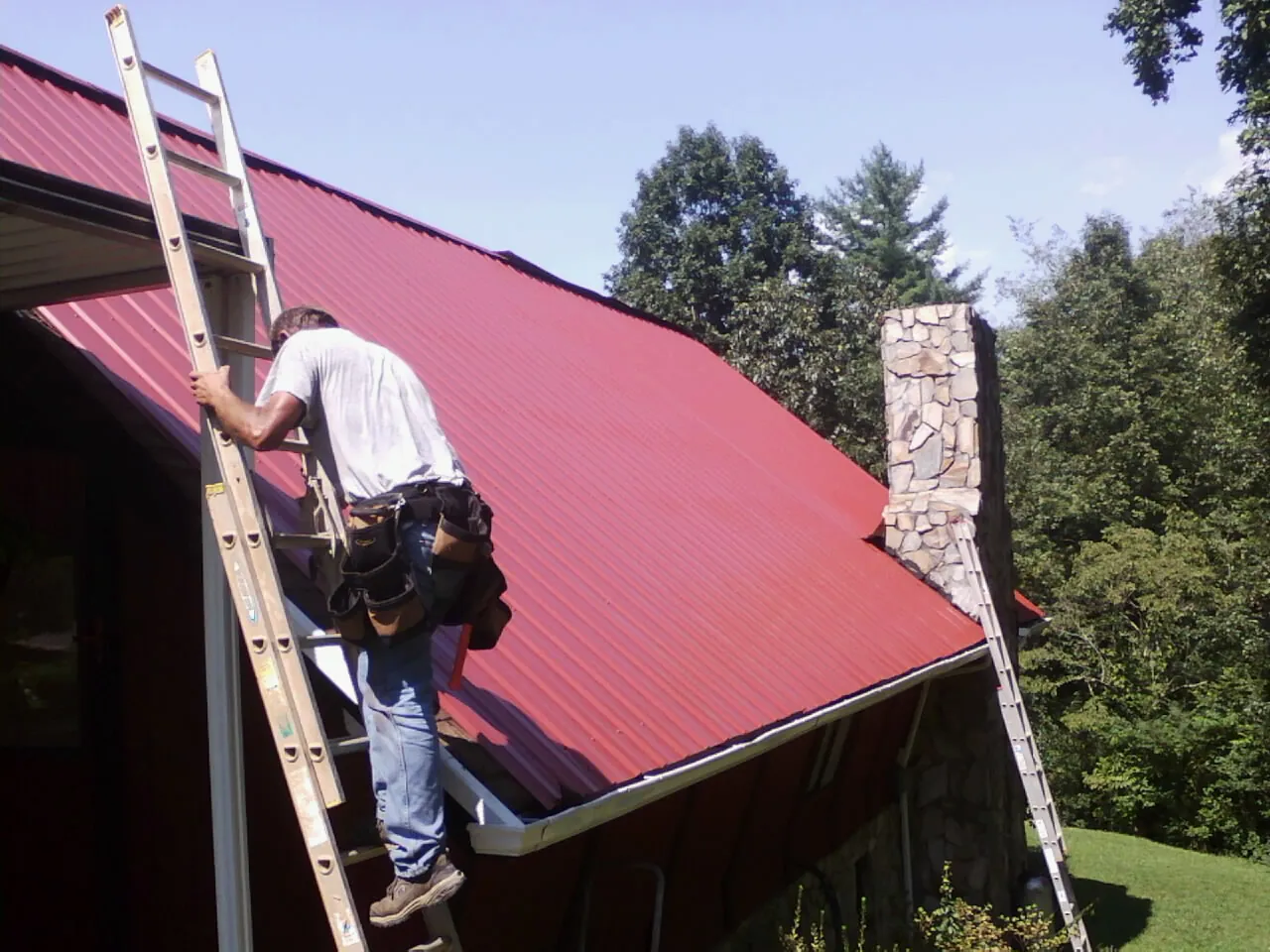 Roofer on a ladder beside a red metal roof and stone chimney — Soffit & Fascia Repair by Roofing Brothers in Garretson